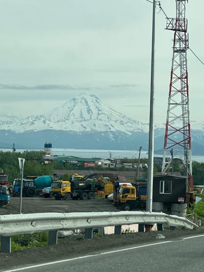 The image captures a panoramic view of the city of Krutoberegovoy, Russia, with a prominent snow-capped mountain range dominating the background. In the foreground, a variety of construction vehicles, including trucks, a cement mixer, and excavators, are parked. A tall communication tower with red and white accents stands to the right of the frame, adjacent to a small, dark utility structure. The scene appears to be an industrial or construction site on an overcast day.