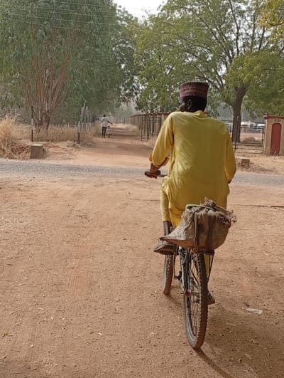 From the back, a man in a yellow robe and a reddish-brown cap is riding a bicycle down a dirt path. The bicycle has a large sack or bag tied to the rear carrier. In the distance, another person is walking and pushing a smaller bicycle in the same direction. Trees line both sides of the path and the sky is lightly clouded. A building is visible on the right of the path near the rider.