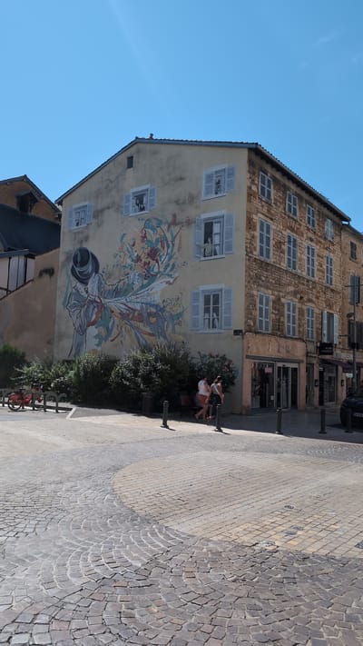 The image depicts a corner building in Villefranche-sur-Saône, France, adorned with a large mural on its pale yellow facade. The mural features a stylized figure in a hat, appearing to blow a trumpet or a similar instrument, with colorful abstract shapes and a procession of smaller figures emanating from it. Several windows with light blue shutters are visible on the building's facade, some with painted figures peering out. To the right, a section of the building is constructed of stone. Below, a street with cobblestone paving curves in the foreground. A few people are seated on a bench in front of the mural, and two individuals are walking past. A red bicycle is parked on the left side of the image. The sky is clear and blue.