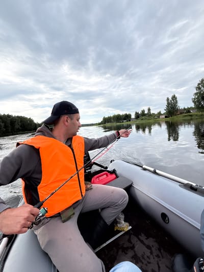 A man in a grey hoodie and an orange life vest sits in a grey inflatable boat, fishing on a calm river. He wears a black baseball cap and is holding a fishing rod. The boat is moving, creating ripples on the water. In the background, the riverbank is lined with trees and houses, suggesting a rural or semi-rural setting. The sky is cloudy and overcast. A red fuel can is visible in the boat. The scene appears to be located in or near Anteplevo (Okhotinskoe S/P), Russia.