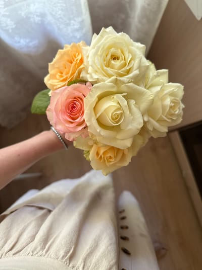 A hand is holding a bouquet of roses. The roses are a mix of cream, pale yellow, peach, and pink. The hand is adorned with a delicate silver bracelet. The bouquet is positioned against a background of a light-colored textured fabric, possibly a dress or drapery, and a wooden surface. In the background, part of a white shoe with a patterned sole is visible, along with the edge of a white piece of furniture that has a lace-like pattern. The lighting suggests it is daytime, and the overall atmosphere is serene. The setting appears to be indoors, with hints of home furnishings. There is no visible text in the image.