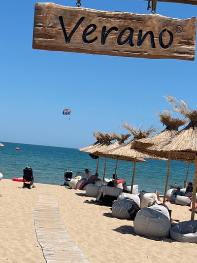 This image depicts a sunny day at a beach in Dyuni, Bulgaria. A wooden sign with the word "Verano" is prominently displayed at the top, suggesting the name of a establishment or location. Below the sign, a beach scene unfolds with the azure sea in the background. Several straw umbrellas provide shade on the sandy shore, under which people are relaxing on beanbag chairs and other seating. A colorful parasail is visible in the sky, indicating recreational activities. A wooden walkway leads across the sand. Strollers are also present on the beach, suggesting families are enjoying the location. The overall atmosphere is one of leisure and relaxation on a summer day.