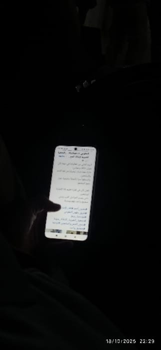 A dimly lit scene in the city of Al-Kiswa, Syria, shows a person's left hand holding a smartphone. The smartphone's screen is illuminated, displaying Arabic text, likely from an article or social media feed, with hashtags visible towards the bottom. Small content thumbnails are present at the bottom of the screen. The individual's thumb is positioned on the screen, suggesting active interaction with the device. The background is almost entirely dark, providing no additional environmental details. The image is timestamped 18/10/2025 at 22:29.