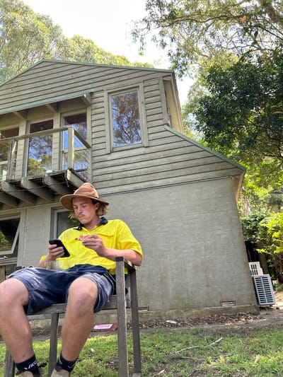 A man in a yellow shirt and brown hat sits on a ladder with a cell phone in one hand and a piece of food in the other. He is in front of a two-story gray house with multiple windows. Trees surround the house, and a gray wall runs along its side. An air conditioning unit sits on top of the wall. The photo is taken on a sunny day.