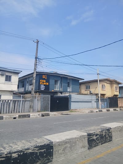 The image depicts a street scene in Animashawun, Nigeria, under a clear blue sky with scattered clouds. In the foreground, a paved road with a black and white striped concrete divider is visible. The divider has a textured, worn appearance. Beyond the road, a series of buildings line the street, separated by fences.
The most prominent building on the left is a light-colored structure with a corrugated metal exterior and a visible air conditioning unit. Adjacent to it is a dark blue building with an orange sign that reads "CK CVR PLACE" with bullet points below listing "SALES", "SERVICE", "INSTALL", and "RENT". This building features balconies and windows, and a utility pole with numerous wires is situated directly in front of it.
To the right of the blue building is a pale yellow house with distinctive arched windows and an air conditioning unit. Further to the right, parts of other structures and fences are visible, including a black metal gate. The overall impression is that of a commercial and residential area in a Nigerian city, with utility infrastructure prominently displayed.