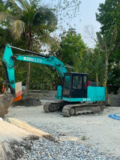 The scene in Hizla, Bangladesh, depicts a stationary teal-colored Kobelco SK05 tracked excavator parked on a light-colored ground composed of sand and gravel. The excavator's boom and bucket are visible on the left, with a piece of white and orange fabric draped over the boom. The cab is black, and the tracks show signs of use. In the immediate foreground, to the left, are piles of light-colored fishing nets with attached weights, resting on a mound of grey aggregate. The background features dense, lush tropical vegetation under a clear sky, including prominent tall palm trees, broad-leafed banana plants, and other green deciduous trees. Stacked concrete blocks are visible behind the excavator. No individuals or active interactions are present in the image.