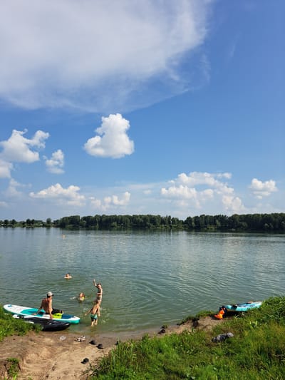 The image depicts a sunny day at a lake, possibly in Sorochii Log, Russia, with a shoreline of sand and grass. The water is calm and reflects the blue sky dotted with fluffy white clouds. In the foreground, on the sandy bank, are several pairs of shoes and a dark-colored garment.

Several people are in the water. Close to the shore, a man wearing a hat sits on a paddleboard. Next to him, a young boy stands in the shallow water. Further out, a girl is in the water, and another person, possibly a child with a yellow flotation device, is also visible. In the distance, a person is in the water with an orange object. Two kayaks or paddleboards are visible near the shoreline on the right side of the image, one is a blue kayak and the other is a life vest.

The background features a dense line of trees along the opposite bank of the lake, with a clear blue sky above. The overall scene suggests a recreational outing on a warm day.