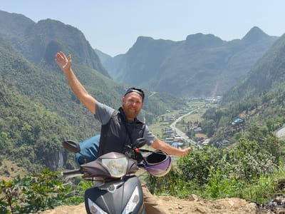 The image captures a fair-skinned man on a scooter with mountains in the background, likely in the Phố Bảng Commune, Vietnam. The man has a beard, wears a black baseball cap, a grey t-shirt, and a dark vest. He is seated on a black and bronze scooter with a blue bag strapped behind him. A purple helmet hangs on the scooter's handlebars. The man is smiling, and his arms are outstretched as if gesturing towards the scenery.
The backdrop consists of mountainous terrain with lush greenery. A valley can be seen with a road and some buildings suggesting a settlement. The sky is light blue. The foreground shows some foliage and a dirt surface where the scooter is parked.