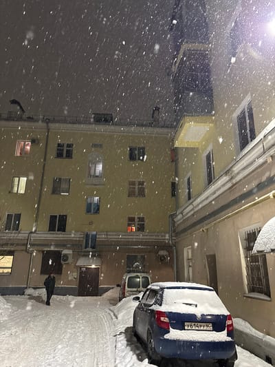 Snow falls heavily in a courtyard surrounded by buildings with illuminated windows. Two cars are parked close to a building, covered in snow. Air conditioning units are visible on the building’s exterior. A figure stands on the snowy ground, possibly looking up at the falling snow. The scene is captured at night with artificial lighting.