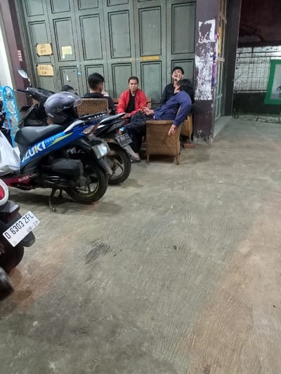 This evening image, set in Cimaung, Indonesia, shows four men gathered casually outside a building with a large, grey folding door, likely a shop or garage. The ground is concrete, and several motorcycles are parked in the foreground.

In the midground, three men are seated on wicker chairs. One man in a red jacket faces forward, while another in a black jacket with a cigarette appears to be laughing or engaged in animated conversation, looking towards the right. A third man, wearing a blue shirt and patterned pants, is slouched back in his chair, appearing relaxed or tired. A fourth man, mostly obscured from behind, sits further to the left.

In the foreground, a prominent blue Suzuki scooter with "SUZUKI" visible on its side has a black helmet resting on its seat. Another dark-colored scooter is parked behind it. Part of a third motorcycle is visible in the extreme foreground, displaying a license plate "D 6303 ZFL" with the date "08-28". A white plastic bag hangs from one of the motorcycles, and a blue plastic crate is visible on the far left.

Attached to the grey folding door are two small signs. The upper sign reads "GAS ADA" (Gas Available), and the lower one reads "BEKAS ADA" (Used/Secondhand Available), indicating the possible nature of the establishment. The artificial lighting suggests it is nighttime or late evening, and the scene depicts a casual social interaction among the men.