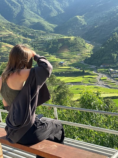 A person is observed from behind, seated on a light brown wooden bench, gazing over a vast mountainous valley in Tú Lệ, Vietnam. The individual has medium-length brown hair and is dressed in an olive green sleeveless top, with a dark grey hooded jacket draped over their shoulders, and dark grey cargo pants. Their right hand is raised near their forehead, and a black smartphone rests on their lap. A grey metal railing and lush green foliage occupy the immediate foreground. The valley below features extensive bright green rice terraces carved into the slopes of verdant mountains. A wide, stony riverbed with visible water channels winds through the valley floor. Scattered small buildings and houses, some with blue roofs, are visible among the terraces and alongside the river. Strong sunlight illuminates the scene, creating bright areas and distinct shadows across the landscape.