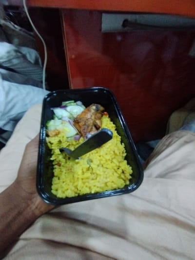 A person is holding a black plastic container filled with yellow rice, a piece of fried chicken, and cucumber slices. The person's right hand is holding the container, while the left hand is holding a black plastic utensil, which is partially submerged in the rice. The container is positioned on the person's lap, and the person is wearing beige pants. The background is blurry, but it appears to be an interior space. The location is Ashaka, Nigeria, with coordinates 10.929, 11.476.