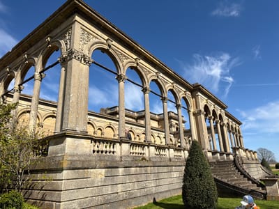 The image depicts an architectural structure in Malvern Hills, United Kingdom. The structure features a series of arches supported by pillars, with intricate decorative details. A balustrade runs along the middle level. Stairs lead up to the structure on the right side. The sky is blue with wispy clouds. The scene is set in a grassy area with a few trees and shrubs, and one person is visible at the bottom right corner of the frame.