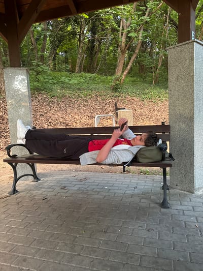 The scene is outdoors under a small shelter with a grey stone top and wooden beams, next to a cobblestone sidewalk. A young person, probably male, sits on a dark-colored wooden bench. He wears a red t-shirt, a grey hoodie, black sweatpants, and white sneakers. He is holding a smartphone and appears to be looking at it. A green backpack sits behind him on the bench. The bench is placed next to a path or road made of cobblestones. In the background, a forest or wooded area is visible.

The image was taken in Gryfino, Poland.