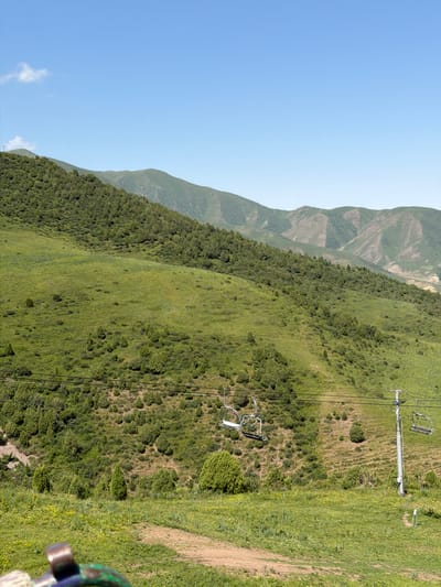 The image depicts a mountainous landscape in Kashka Suu, Kyrgyzstan, under a clear blue sky with a few wispy clouds. In the foreground, a grassy area with scattered yellow wildflowers leads to a dirt path. A portion of a metal railing, painted in green and blue, is visible at the bottom left. Beyond the foreground, the scene is dominated by a lush green mountainside covered in dense, low-lying trees and bushes, with taller trees scattered throughout.

A ski lift runs across the hillside, with several empty chairlifts suspended from cables. One ski lift tower, a tall metallic structure, is clearly visible on the right side of the image. The mountain slopes recede into the background, displaying successive layers of peaks and valleys, some with lighter green vegetation and barren patches. The overall impression is one of a natural, undeveloped mountain environment, likely a recreational area during the summer season. No people or animals are present in the frame.