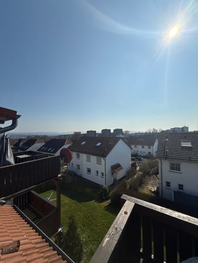 The image depicts an elevated view of a residential neighborhood in Vellmar, Germany, under a clear, bright blue sky with direct sunlight in the upper right quadrant creating a lens flare.
In the immediate foreground, to the left, are terracotta roof tiles, a grey gutter, and a dark brown wooden balcony railing with vertical slats. Below the gutter, a dark, possibly tarp-covered object is visible. To the right, another section of a dark wooden balcony railing, showing some wear, frames the scene.
The midground features several detached residential houses with white facades and dark, gabled roofs. The most prominent house, centrally located, has a red satellite dish mounted on its roof and multiple visible windows. This house also features a small, covered entryway on its side and a well-maintained green lawn in its backyard. Other similar houses are visible beyond it, some with skylights on their roofs.
In the background, a distant cityscape with numerous multi-story buildings, likely apartment blocks or commercial structures, is visible on the horizon, indicating a larger urban area beyond the immediate residential zone. Subtle hills are faintly discernible even further in the distance. Vegetation includes green grass, some bare trees typical of cooler seasons, and evergreen shrubs around the houses. No individuals are visible in the scene. The overall impression is a sunny, quiet day in a suburban setting.