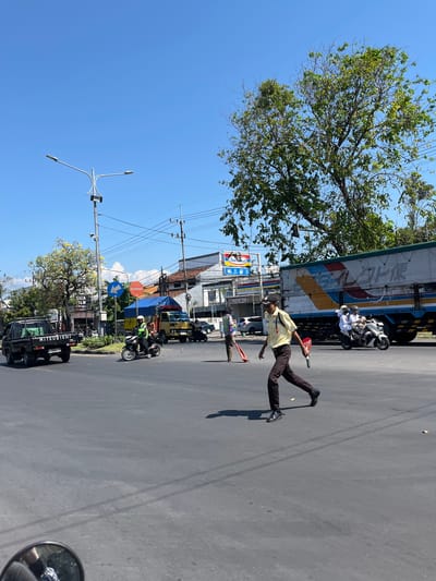 This is a street-level, eye-level, daytime, medium shot of a busy intersection in Klurak, Indonesia. The scene is dominated by a paved intersection under a clear blue sky. In the foreground, a security guard in a yellow short-sleeved shirt, brown pants, a black cap, and black shoes is walking across the intersection, holding a red stop sign in his right hand. He is looking towards the right. Behind him, a white motorcycle with two occupants wearing white clothing and helmets is passing. To the left, a black Mitsubishi pickup truck is parked. Further in the background, a large blue and white truck is stopped at the intersection, with a blue tarp covering its cargo. To the left of the truck, a yellow truck is also visible. Various buildings, including a shop with "Indomaret" signage, line the street. A tall street lamp with multiple lights stands on the left side of the intersection. Power lines crisscross the sky. Several trees, one with yellow flowers, are scattered throughout the scene. The overall atmosphere is that of an active urban environment during daylight hours.