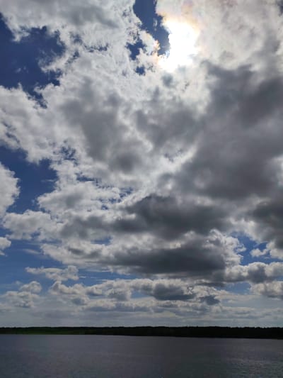 The image depicts a landscape with a body of water, possibly a lake, in the foreground, a line of trees along the horizon, and a sky filled with clouds. The sky is mostly cloudy with some patches of blue visible. The sun is present, partially obscured by the clouds, creating a bright spot in the sky.
While the image strongly suggests a natural setting with a lake and trees, it does not contain any distinct features that allow for a definitive location within the city of Pruzhinino, Russia. Lacking any information about visible buildings, landmarks, or other contextual elements in the image. The given information limits the ability to confirm the location without additional information.
