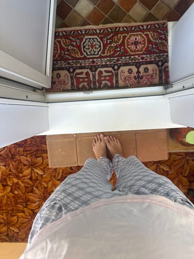 A first-person perspective shot shows a person standing in what appears to be a doorway. The floor is covered with two different rugs. The rug closer to the viewer has a large floral pattern in shades of orange and brown. Beyond that, there is a smaller rug with a traditional geometric design in red, black, and beige. The doorway itself is framed by white trim. The person is wearing plaid pajama bottoms. Their bare feet are visible on a small section of terracotta-colored tile that bridges the gap between the rugs and the doorway. To the right of the frame, a large glass jar with a green lid, possibly containing tomatoes, rests on the floor. The scene is indoors, likely in a residential setting. The context of Rasskazovo, Russia, is not visually evident in the image itself.
