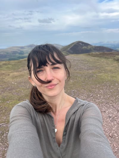 The image is a selfie taken outdoors on a hilly, barren landscape. The subject is a woman with dark hair and bangs, wearing a grey cardigan and a silver necklace with a heart-shaped pendant. She is smiling and looking towards the camera. Her hair is blowing across her face. In the background, there are rolling hills under a cloudy sky. The ground is covered in gravel and sparse vegetation. The overall scene suggests a hike or outdoor excursion in a rural location. The specific location in Penicuik, United Kingdom cannot be determined from the image alone.
