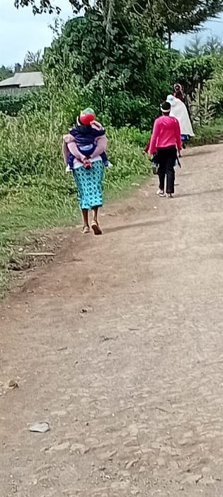 In Gatakaine, Kenya, an unpaved dirt path extends from the foreground into the background. Three individuals are observed walking away from the camera. The leftmost person wears a light-colored top, a patterned blue and white skirt, and light-colored sandals, carrying a child secured on their back. The child wears a red cap or head covering. The middle individual wears a long-sleeved pink shirt, dark trousers, and a dark head covering with a white band, carrying an unidentified dark object in their right hand. The third individual, positioned to the right of the second and partially obscured, wears a white garment that covers their body and head. To the left of the path, dense green vegetation, comprising tall grasses, bushes, and trees, lines the route. A single-story building with a light-colored roof is visible in the upper-left background, partially obscured by the foliage. The sky above is overcast.