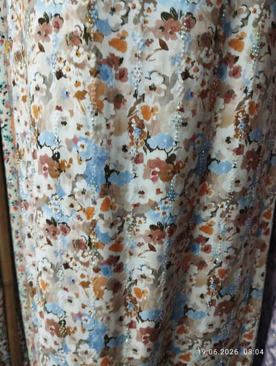 The image shows a close-up of fabric with a floral print and scattered embellishments. The print features various flowers in shades of white, orange, brown, and blue against a light beige background. Vertical lines of small, iridescent beads or sequins run through the fabric, adding a sparkling effect. The fabric appears to be draped, creating folds and shadows that give it texture. In the bottom right corner, a timestamp is visible: "19.06.2026 08:04". The scene is forensic and matter-of-fact, describing the visual elements without inferring narrative or context beyond what is presented. No people or specific environmental details outside of the fabric itself are discernible.