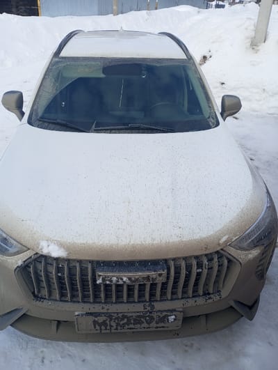 This image presents the front view of a white crossover SUV, extensively covered in dark brown dirt and mud, situated outdoors in the city of Yaromaska, Russia. The vehicle's hood, large dark grille, and front bumper are heavily soiled, with some patches of snow visible on the grille and in front of the bumper. The area designated for the license plate is completely obscured by dirt. The windshield shows a scattering of dirt, with two wipers in their standard resting position; the interior reveals a steering wheel and rearview mirror. The vehicle is equipped with black roof rails. The ground surrounding the SUV is uniformly covered with white snow. The background consists of substantial snowdrifts, a light-colored corrugated metal fence, and vague, distant structures. No individuals are present, and no actions or interactions are depicted.