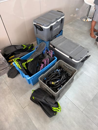 The image shows a collection of diving equipment arranged on a gray tiled floor. Two gray hard cases, one labeled "ICEMASTER", rest on a blue plastic crate. Next to them, a gray plastic crate filled with black hoses or cables sits atop another blue plastic crate. These crates are filled with black and purple dive fins. Some black fins with yellow accents are also visible outside the blue crate. The scene appears to be indoors. In the upper left corner, a person's bare feet are visible near a white and chrome stool.