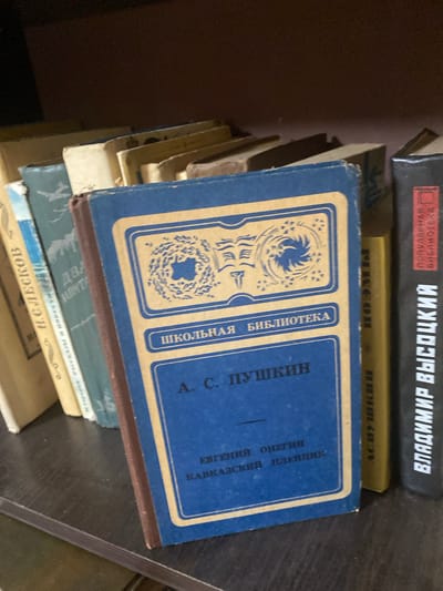 The image shows a bookshelf with several books. The main focus is a blue hardcover book with gold lettering and decorative elements on the cover. The title on the book appears to be "А. С. ПУШКИН" and below that, "ЕВГЕНИЙ ОНЕГИН" and "КАВКАЗСКИЙ ПЛЕННИК". Other books are visible on the shelf, some with their spines facing forward, displaying text in Cyrillic. One book on the right has "ВЛАДИМИР ВЫСОЦКИЙ" written vertically on its spine. The bookshelf is dark brown. The scene is static, with no people or animals present. The lighting suggests an indoor environment, possibly a home or library. The location is identified as Афанасьево, Russia, but this context is not visually evident from the image itself.