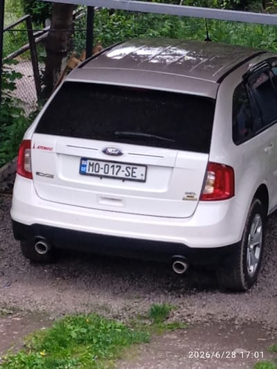 The image shows the rear view of a white Ford Edge parked outdoors. The car has a license plate with the text "GE MO-017-SE". The background features lush green foliage and trees, suggesting a natural or rural setting. A chain-link fence is partially visible in the upper left corner. The car is positioned on a gravelly surface with some patches of grass. The overall lighting and the presence of dew or water droplets on the car's roof suggest it might be early morning or that it has recently rained. The timestamp "2026/6/28 17:01" is overlaid on the bottom right of the image, indicating the date and time the photo was taken. The scene appears to be a quiet, daytime snapshot of a vehicle in a semi-natural environment. There are no people visible in the image.