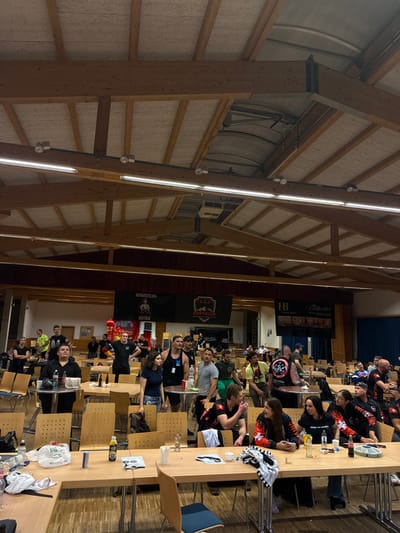 This image depicts an indoor event within a large hall located in Reisenberg, Austria. The venue features a high, vaulted wooden ceiling with exposed structural beams and white acoustic paneling, illuminated by multiple linear fluorescent light fixtures and smaller track lights mounted on horizontal bars. The floor is wooden.
Approximately 30-40 adult individuals are visible, standing and seated at numerous light-colored wooden folding tables (both rectangular and round) and matching chairs arranged throughout the hall. Many individuals are engaged in conversation, and various bottles, cans, and cups containing beverages are present on the tables, alongside other miscellaneous items and personal belongings.
In the left foreground, a woman in a black top is seated at a small round table. In the midground, several men, some with noticeable muscular physiques, are standing and conversing. In the lower right foreground, a group of women, many wearing black tops with red accents or branding, are seated at a rectangular table, interacting socially. Most attendees are dressed in casual attire.
In the background, three large banners are affixed to the far wall: the left banner displays "ARM WRESTLING AUSTRIA" with a graphic of two arm wrestlers; the central banner features the "A.A.E. AUSTRIAN" eagle logo; and a partially obscured banner to the right reads "HB HOTEL REISINGER." A prominent red inflatable object is also visible in the left background. The scene suggests a social gathering, possibly a post-competition or break area for an arm-wrestling event.