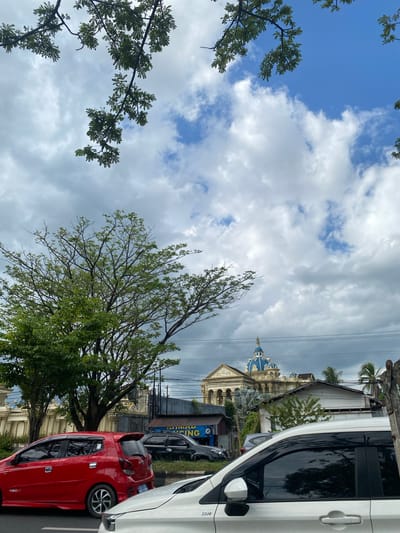 A street scene in Mandar Sari, Indonesia, is captured with a low-angle perspective, looking upwards towards a sky filled with clouds. Two cars are parked on the right side of the image: a white car in the foreground, and a red car behind it. A dark-colored vehicle is parked further down the street. The white car's side, including its door handle and side mirror, is prominently visible. The red car's rear section and wheel are also in view.
In the background, several buildings are visible, with one notable structure featuring a distinctive blue dome. Trees with green foliage are interspersed throughout the scene, some framing the sky and others partially obscuring the buildings. Power lines stretch across the upper portion of the image, adding to the urban feel. The weather appears to be overcast, with a mix of bright and dark clouds suggesting a partly cloudy or changeable day. The time of day is not explicitly indicated but the lighting suggests it could be daytime. The overall mood is calm and observational, depicting a typical street view. A partial sign with the text "SING" is visible on a building in the background.
