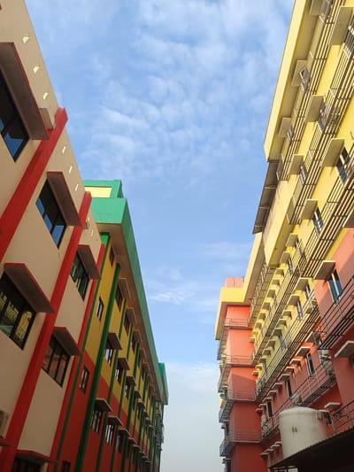 The image shows a low-angle view of the exterior of two brightly colored buildings, one on the left and one on the right, against a bright blue sky with scattered white clouds. The building on the left is cream-colored with red and green accents and multiple windows. The building on the right is primarily yellow with red and pink sections, also featuring numerous windows and metal railings. The buildings appear to be modern, possibly an educational institution or office complex, located in the city of Karangtengah, Indonesia. The scene is devoid of people, capturing only the architectural elements and the sky. The lighting suggests it is daytime, with the sun casting shadows that emphasize the architectural details.