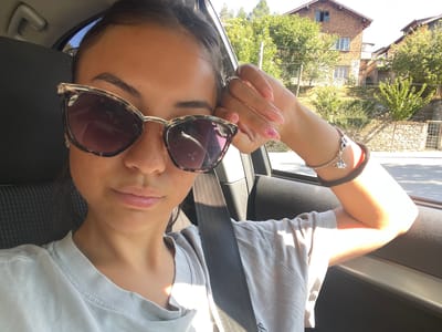 A young woman with dark hair pulled back is taking a selfie inside a car. She is wearing sunglasses with a tortoiseshell frame and a light gray t-shirt. Sunlight streams in from the right, illuminating her face and arm. She rests her right hand on her face, showing off pink nails with a floral design and a silver ring. A black seatbelt is visible across her chest. Outside the car window, a village scene is visible with traditional brick houses and lush green trees. The woman appears to be in Belovo, Bulgaria, judging by the architecture and vegetation.