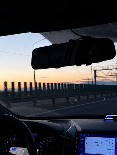 The image shows a view from inside a car, likely on a highway or bridge. The rearview mirror is prominently displayed in the upper left of the frame. Through the windshield, a highway with repetitive structures like overpasses or railings can be seen, rendered blurry by motion. The scene suggests it's either dawn or dusk due to the warm colors of the sky. The car's dashboard is visible, including a speedometer indicating a slow speed or standstill and a multimedia display showing a map. The map shows a location near Kislyakovskaya, Russia. The map display on the infotainment system indicates the car's location is 71 km from the final destination, and the date displayed is May 11, 2026.