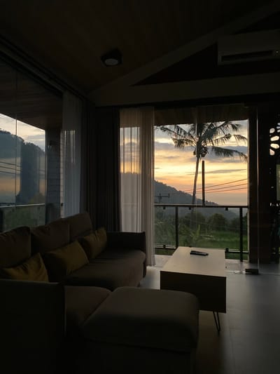 The image depicts an indoor living space with a large window offering a view of a tropical sunset. A sectional sofa is positioned in the foreground, facing the window. A coffee table with a remote control on it sits in front of the sofa. Through the window, a palm tree is prominent against a sky filled with the warm colors of sunset, including orange, yellow, and pink. In the distance, rolling hills and lush vegetation are visible, suggesting a rural or elevated location. The lighting indicates it is dusk. The scene is likely a private residence or a resort accommodation in Sawan, Indonesia, given the tropical elements and architectural style.
