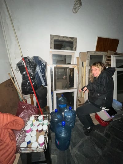An image, rotated 90 degrees counter-clockwise, depicts a young woman posing in a cluttered indoor or semi-enclosed space, likely a storage area or workshop in Saladillo, Argentina. The scene is dimly lit, suggesting nighttime, with artificial light highlighting the subjects.

The woman, with light-colored hair, wears a dark jacket and dark pants, and is kneeling on a dark, possibly tiled or stone floor. She smiles widely at the camera, conveying a cheerful and playful expression. Her body is positioned next to a vertical stack of various window frames, some containing glass, leaning against a white wall. The frames appear to be of different styles and conditions.

Numerous objects fill the foreground and midground. To the woman's right, several large, blue plastic water dispenser bottles are stacked, with the brand name "Quillagua" visible on one. Below these bottles, a metal wire basket holds many small jars with colorful lids (pink, green, white, blue), suggesting craft supplies or cosmetics. Nearby, a black mannequin-like torso is visible, along with various plastic-wrapped items (possibly clothing), red plastic bags, and long poles or sticks. A red plastic basket is also visible near the woman's lap, while a brown wooden board leans against a wall in the background. The surrounding walls are light-colored, showing some marks and textures.

The overall setting suggests a utilitarian or storage area, perhaps undergoing renovation or used for various projects, typical of a workshop or storage room.