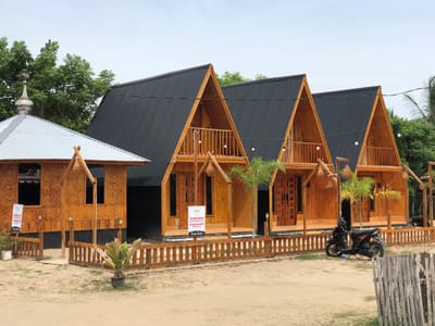 This media file depicts a row of three identically designed A-frame wooden cabins with black corrugated metal roofs, set against a backdrop of lush green trees under a bright, overcast sky. To the left of the cabins is a smaller, single-story wooden structure with a silver corrugated metal roof and a decorative dome, resembling a small mosque or pavilion. The cabins and the smaller structure are constructed with prominent wooden beams and natural wood paneling, giving them a rustic and tropical feel.
In front of the cabins, a light brown wooden fence encloses the area. A few potted plants and some dry vegetation are visible near the fence and the cabins. A dark-colored motorcycle is parked to the right of the cabins, near the fence. The ground in front of the cabins appears to be sandy or dirt, suggesting a beach or rural setting. Hanging light bulbs are strung between the cabins, possibly for evening illumination.
Visible text on a white sign in front of the leftmost cabin reads "DILARANG MASUK SELAIN TAMU VILLA DILARANG MELOMPAT PAGAR VILLA Terima Kasih" which translates to "NO ENTRY OTHER THAN VILLA GUESTS. DO NOT JUMP THE VILLA FENCE. Thank you." Another sign on the far left cabin has some unreadable text.
The overall scene suggests a resort or guesthouse in a tropical location, likely during the daytime, given the bright sky, although the overcast conditions suggest it might not be midday with direct sunlight. The setting appears peaceful and welcoming, with the architecture emphasizing natural materials and a connection to the environment.