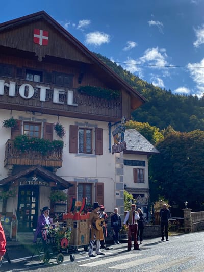 The image depicts a street scene in Abondance, France, featuring the Hôtel Les Touristes on a sunny day. The prominent building, identified by large "HOTEL" lettering and a hanging sign reading "Les Touristes HÔTEL & RESTAURANT," is a multi-story structure with a brown wooden gable roof adorned with a red shield bearing a white cross (Swiss flag emblem). Balconies with decorative wooden railings and flower boxes containing pink flowers are visible on the upper levels. The lower facade is light-colored with stone corner accents and windows with dark brown shutters. An awning over an entrance also reads "Les Touristès."
On the asphalt road in front of the hotel, a group of five individuals is present, including three musicians and two onlookers, positioned near a pedestrian crossing. One musician on the left wears a purple long coat and a black top hat, operating an organ grinder on a two-wheeled cart. Another musician, dressed in a brown long coat, black and white striped trousers, and a black top hat, plays an acoustic guitar next to a large, elaborate wooden musical prop with red accents. A third musician, wearing a red and white striped costume with a vest and a red hat, holds a microphone. Two individuals observe the performance from behind the musicians on the right side of the road.
In the background, a dense forest covers a mountain under a blue sky with scattered white clouds. To the immediate right of the main hotel building, an adjacent structure with a grey sloped roof and similar architectural features is visible. A low stone wall lines the right side of the road.