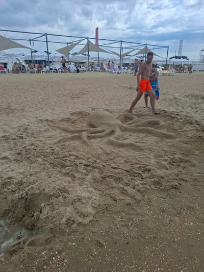The image depicts a sandy beach in Vityazevo, Russia, under a cloudy sky. In the foreground, a sand sculpture of an octopus is prominent, with its tentacles spread across the sand. Two people, a man wearing orange shorts and a child wrapped in a blue and white towel, are standing near the sculpture. The man is looking towards the sculpture. In the background, a beach resort area is visible with rows of lounge chairs, umbrellas, and beach tents, suggesting a public recreational space. Several people are seated or walking amongst the beach amenities. A tall white structure with some signage is also present on the far right. The sand is damp in some areas, particularly near the water's edge, and scattered with shells.