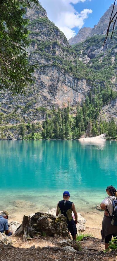 The image depicts a stunning natural landscape with a focus on a vibrant turquoise lake bordered by a steep, verdant mountain. Two people are visible in the foreground, seated on a rocky outcrop overlooking the water. One individual wears a black bandana and a light-colored top, with a backpack on. The other person is wearing a blue cap and a dark, sleeveless shirt with shorts. Another person is partially visible in the lower right corner, wearing a blue shirt and a hat. The scene suggests a leisurely moment of admiration for the breathtaking scenery.

The mountain slope is densely covered with trees, creating a striking contrast with the sheer rock faces and the clear, inviting water. The sky above the mountains appears blue with scattered white clouds, indicating fair weather. The time of day appears to be daytime, likely late morning or afternoon, given the lighting conditions. The location cues, such as the style of the mountains and the characteristic turquoise color of the water, are consistent with the Prags - Braies region of Italy. There is no visible text in the image.