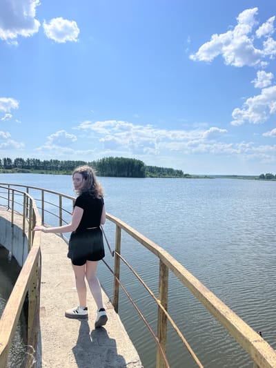 A young person is on a bridge over water, possibly a canal or river. They are wearing a black short-sleeved shirt, a black skirt, and white sneakers with black stripes. The person is leaning forward, with their right hand touching the railing of the bridge. The bridge has a yellow rusted metal railing and a concrete surface. The water is calm with small ripples. Trees line the opposite bank of the water. Above, there is a blue sky with scattered white clouds. The location is Korovino, Russia.