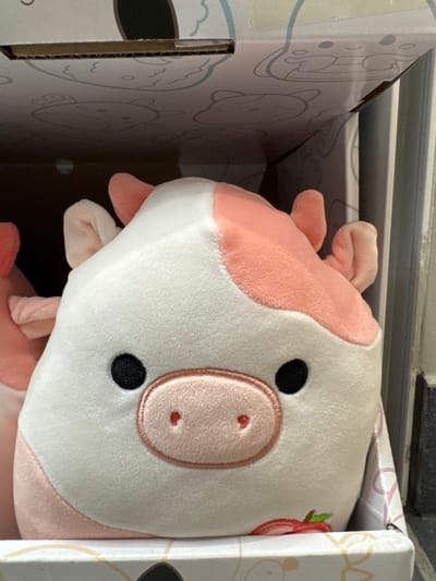 The image shows a close-up of a plush toy, likely a cow, peeking out of a cardboard box. The toy is predominantly white with pink patches and features. It has a round snout with embroidered nostrils, black eyes, and small pink ears. A hint of a red strawberry design is visible on the right side of the toy. The box has a light color with a subtle pattern of cartoon animals, including what appears to be a bear. The background is indistinct, suggesting an indoor setting. The lighting appears to be natural, coming from above and to the left, casting soft shadows. The image focuses on the toy and the edge of the box, with the rest of the box interior and the surrounding environment out of focus. There is no visible text. The overall impression is one of a new item, possibly being unpacked.