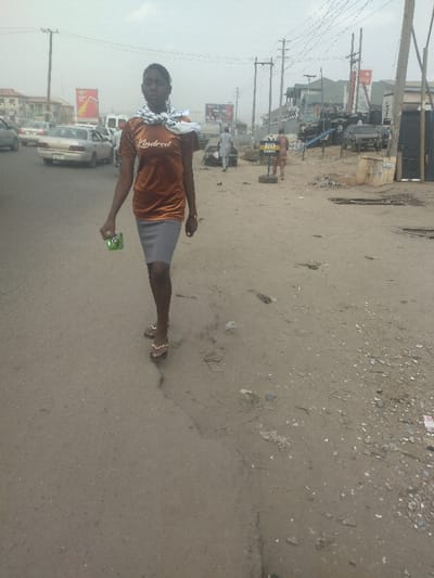 A young woman in a brown shirt with the word "Indred" printed on the front and a gray skirt is walking on a dirt road. She is holding a green object in her left hand. She is walking towards the camera and her face is visible. There are other people in the distance, including a man in a red shirt and a white shirt. The road is dusty and has a crack down the middle. There are cars parked on either side of the road. There is a sign in the distance that says "No Parking." The location is in the city of Way, Nigeria, at latitude 9.029 and longitude 7.584.