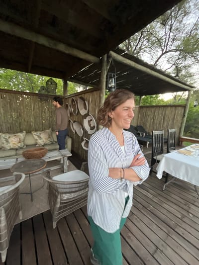 The image depicts an outdoor setting, potentially a patio or deck, possibly located in the vicinity of Khwai, Botswana. A woman in a blue and white striped shirt and green pants stands with her arms crossed, smiling. There are wooden planks, a table with a white tablecloth and place settings, and several chairs in the background. In another seating area, there is a person in a brown shirt lying down. The area is decorated with what appears to be basket-like items and cushions. The architecture is somewhat rustic, with wooden support beams and what seems like a thatched roof. The environment suggests a safari lodge or similar establishment in a natural setting.