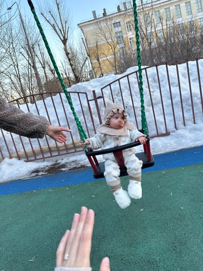 A baby wearing a beige knitted hat, matching sweater with small heart patterns, and fuzzy white boots is seated in a red and black swing. A brown, possibly edible or pacifier-like, object is in the baby's mouth. An adult's arm, clothed in a brown textured sleeve, is visible reaching towards the baby from the left. In the lower foreground, a human hand with a silver-colored ring on the ring finger is visible, out of focus. The swing hangs over a green and blue rubberized playground surface. In the background, large piles of snow are present behind a brown metal fence, along with several bare deciduous trees. A multi-story yellow building with white windows is visible in the distant background under a clear blue sky. The scene takes place outdoors in Sadovaya, Russia, under natural daylight conditions.