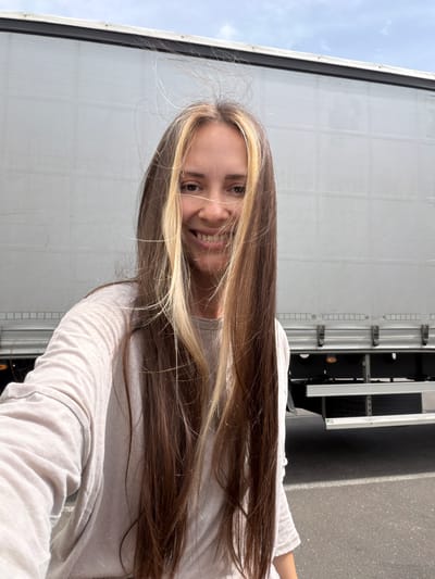 A woman is standing next to a truck. The location is the city of Sluchainyi, Russia. The woman has long brown hair with blonde highlights and is wearing a light-colored long-sleeved shirt. She is smiling. The truck is white with black components. The background includes a street or parking area and a cloudy sky.