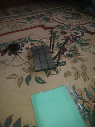 A black wireless router with three antennas and a black power adapter are placed on a patterned carpet. A teal colored paper with handwritten text is also on the carpet, along with a clear plastic sleeve. The text on the paper includes a list of numbered items with letters, an email address, and a phone number. The scene appears to be indoors, possibly in a residential setting in Jıbek joly, Kazakhstan.