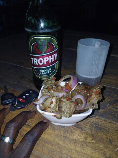 A photograph, possibly taken indoors, depicts a Trophy brand beer bottle lying horizontally on a wooden surface. Next to the bottle is a white bowl containing what appears to be spicy food, possibly offal, mixed with red onion slices. A translucent plastic cup sits next to the bowl. A hand, with a silver ring visible on one of the fingers, is also present. A car key is on the wooden surface near the hand and bowl. The specific location within Kagwaru, Nigeria cannot be determined from the image. No other people, actions, or interactions are visible in the image.
