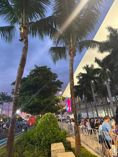 An outdoor evening scene at an event in Miami Gardens, United States of America, features a crowd of people lining up behind a white barrier. Palm trees are prominent throughout the image, with two large palm trees in the foreground. In the background, a large, modern white building with a curved roof is visible, possibly a stadium or arena, with a large digital screen displaying colorful advertisements. Several more palm trees line the front of the building. The sky is a dusky purple and blue, indicating it is either dawn or dusk. A red Coca-Cola tent is situated in the middle ground, with a blue recycling bin next to it. The people in the crowd appear to be dressed casually, and some are looking towards the building or the screen. Some are holding what appear to be tickets or bags. The overall atmosphere suggests an organized event with people anticipating entry or an activity.